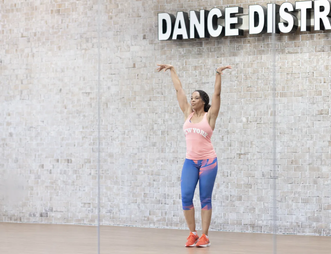 Sophie in a dance studio, both arms raised overhead, beneath a DANCE DISTRICT sign.